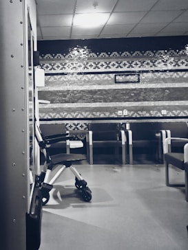 A clinical waiting area with a row of empty chairs against a wall decorated with geometric patterns. A wheelchair is parked to one side, and the lighting is soft and subdued. The setting seems orderly and quiet with a sterile ambiance.
