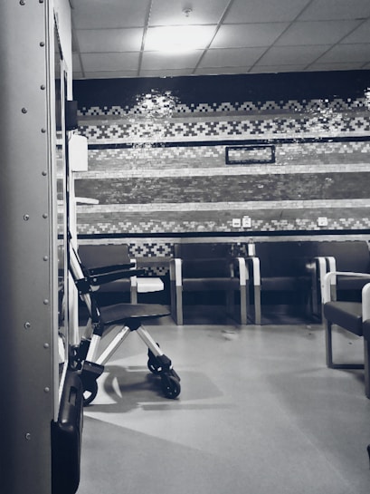 A clinical waiting area with a row of empty chairs against a wall decorated with geometric patterns. A wheelchair is parked to one side, and the lighting is soft and subdued. The setting seems orderly and quiet with a sterile ambiance.