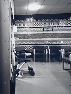 A clinical waiting area with a row of empty chairs against a wall decorated with geometric patterns. A wheelchair is parked to one side, and the lighting is soft and subdued. The setting seems orderly and quiet with a sterile ambiance.