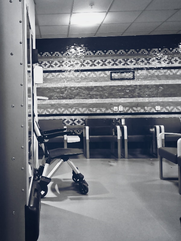 A clinical waiting area with a row of empty chairs against a wall decorated with geometric patterns. A wheelchair is parked to one side, and the lighting is soft and subdued. The setting seems orderly and quiet with a sterile ambiance.