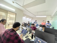 A group of people in an office setting working on laptops, with several individuals engaged in conversation. The room is well-lit with fluorescent lights and has glass partitions.