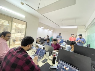 A group of people in an office setting working on laptops, with several individuals engaged in conversation. The room is well-lit with fluorescent lights and has glass partitions.