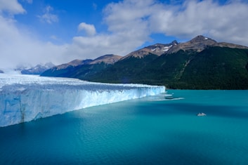 A vast glacier extends into a turquoise body of water, with a small boat visible on the water's surface. The glacier is framed by rugged mountains with some vegetation visible on their slopes. The sky above is partly cloudy, adding depth to the dramatic landscape.