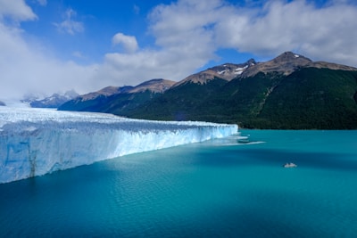 A vast glacier extends into a turquoise body of water, with a small boat visible on the water's surface. The glacier is framed by rugged mountains with some vegetation visible on their slopes. The sky above is partly cloudy, adding depth to the dramatic landscape.
