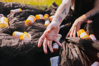 A hand reaching out over a pile of orange prescription pill bottles scattered on a black fabric. The arm has visible tattoos, and there are grassy surroundings suggesting an outdoor area.