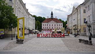 A cobblestone plaza leading to a historic building with a central clock tower, flanked by symmetrical rows of classical structures. In the foreground, a large red hashtag sculpture reading '#TARTU2024' is prominently displayed, accompanied by a large yellow rectangular frame on the left. Flags and outdoor seating with umbrellas are visible, suggesting a lively and inviting atmosphere.