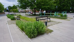 A landscaped urban area featuring paved walkways and benches surrounded by green plants. There are several electric scooters parked in designated areas, and trees lining the street. Cars are parked along the road, and buildings can be seen in the background.