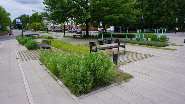 A landscaped urban area featuring paved walkways and benches surrounded by green plants. There are several electric scooters parked in designated areas, and trees lining the street. Cars are parked along the road, and buildings can be seen in the background.