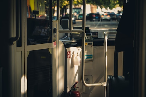 Interior view of a bus showing the driver's cabin and entrance area with a focus on the fare collection machine. Sunlight streams through windows, casting shadows on the interior surfaces. A fire extinguisher is visible near the door.