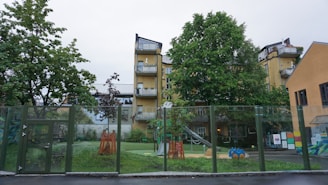 A playground is surrounded by a glass fence with various toys and equipment, including a slide and a blue toy car. There are trees providing greenery, and behind them stands a multi-story yellow building with balconies. The setting appears calm and residential.