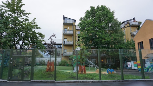 A playground is surrounded by a glass fence with various toys and equipment, including a slide and a blue toy car. There are trees providing greenery, and behind them stands a multi-story yellow building with balconies. The setting appears calm and residential.