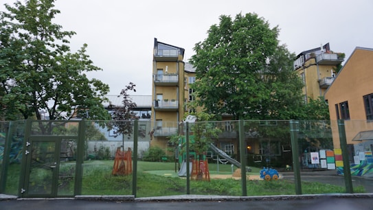 A playground is surrounded by a glass fence with various toys and equipment, including a slide and a blue toy car. There are trees providing greenery, and behind them stands a multi-story yellow building with balconies. The setting appears calm and residential.