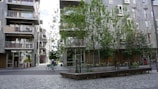 Urban scene featuring a street with modern apartment buildings and trees. A person is riding a bicycle across the paved area. The buildings have balconies and large windows, and there are several benches under the trees.