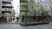 Urban scene featuring a street with modern apartment buildings and trees. A person is riding a bicycle across the paved area. The buildings have balconies and large windows, and there are several benches under the trees.