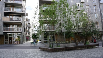Urban scene featuring a street with modern apartment buildings and trees. A person is riding a bicycle across the paved area. The buildings have balconies and large windows, and there are several benches under the trees.