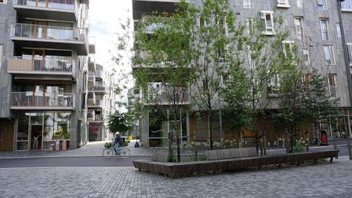 Urban scene featuring a street with modern apartment buildings and trees. A person is riding a bicycle across the paved area. The buildings have balconies and large windows, and there are several benches under the trees.