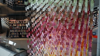 A library setting features cascading letters in various colors, suspended decoratively from the ceiling. Bookshelves filled with books line the walls, and a person is visible near the lower center of the image next to an escalator.