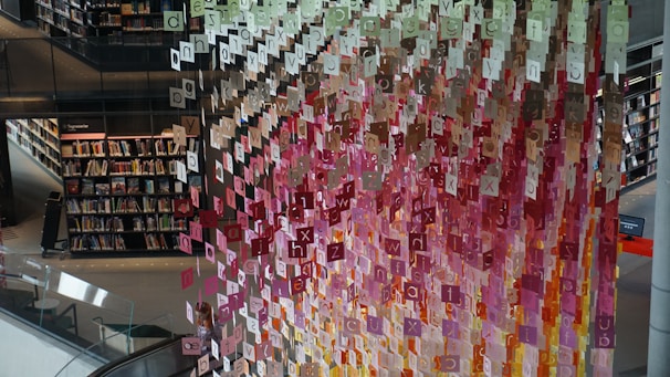 A library setting features cascading letters in various colors, suspended decoratively from the ceiling. Bookshelves filled with books line the walls, and a person is visible near the lower center of the image next to an escalator.