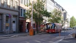 A red public bus is traveling down a quiet urban street lined with tall buildings and green trees. There is a barrier in a raised position near the curb, and various street signs are visible. The streets are clean and the overall environment appears peaceful.