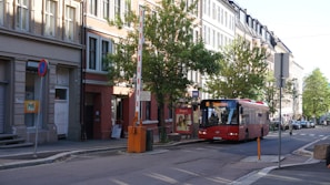 A red public bus is traveling down a quiet urban street lined with tall buildings and green trees. There is a barrier in a raised position near the curb, and various street signs are visible. The streets are clean and the overall environment appears peaceful.