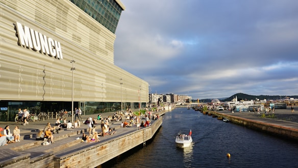 A modern art museum with the word 'MUNCH' displayed prominently on a large, contemporary building. There are people relaxing and socializing on a wooden deck next to a canal where a small boat is floating. In the background, urban buildings and a cloudy sky are visible.