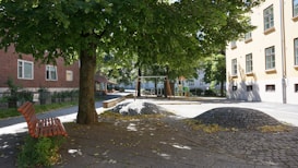 A peaceful urban park scene featuring a tree providing shade over a wooden bench. The background includes a playground with swings and a slide. Two buildings with windows can be seen, one with red brick and the other cream-colored. The ground has cobblestone areas with leaves scattered around, indicating a serene setting.