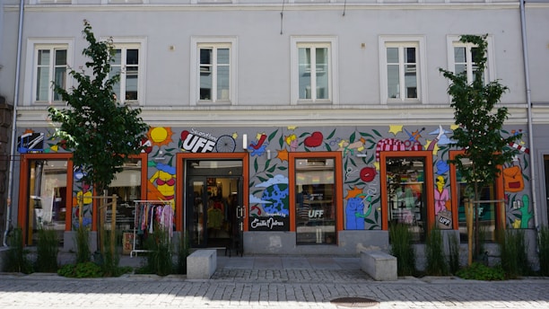A storefront with colorful wall art including abstract shapes, lips, fruit, stars, and various designs. The building is two stories tall with several windows and trees planted in front. Clothes are visible inside the glass storefront.