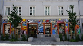 A storefront with colorful wall art including abstract shapes, lips, fruit, stars, and various designs. The building is two stories tall with several windows and trees planted in front. Clothes are visible inside the glass storefront.