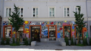 A storefront with colorful wall art including abstract shapes, lips, fruit, stars, and various designs. The building is two stories tall with several windows and trees planted in front. Clothes are visible inside the glass storefront.