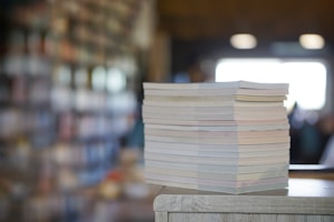 A stack of books or magazines is placed on a wooden surface, with a blurred background of bookshelves filled with various items. The scene suggests a library or bookstore environment with soft natural lighting coming from a window.