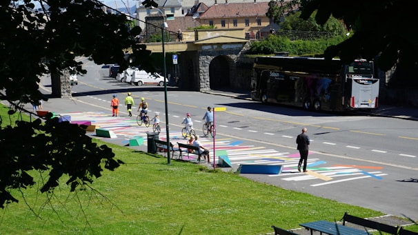 A street scene featuring a colorful painted crosswalk where several people are riding bicycles, and a few pedestrians are present. A couple of individuals wearing bright safety clothing stand on the sidewalk, observing. Parked vehicles and a large bus are visible on the side of the road. The scene is bordered by greenery, and a stone bridge is in the background.