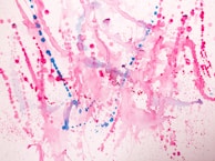 Vibrant splashes of pink and blue paint create an abstract pattern on a white background. The composition consists of various dots, splatters, and fluid lines, with each color blending and overlapping in places. The overall effect is energetic and playful, with a sense of dynamic motion capture.