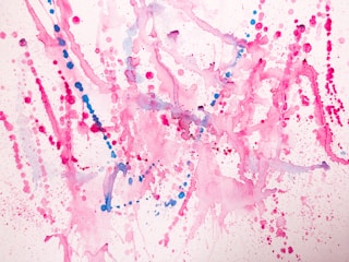 Vibrant splashes of pink and blue paint create an abstract pattern on a white background. The composition consists of various dots, splatters, and fluid lines, with each color blending and overlapping in places. The overall effect is energetic and playful, with a sense of dynamic motion capture.