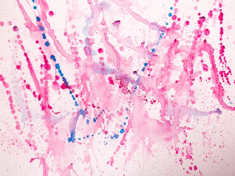 Vibrant splashes of pink and blue paint create an abstract pattern on a white background. The composition consists of various dots, splatters, and fluid lines, with each color blending and overlapping in places. The overall effect is energetic and playful, with a sense of dynamic motion capture.