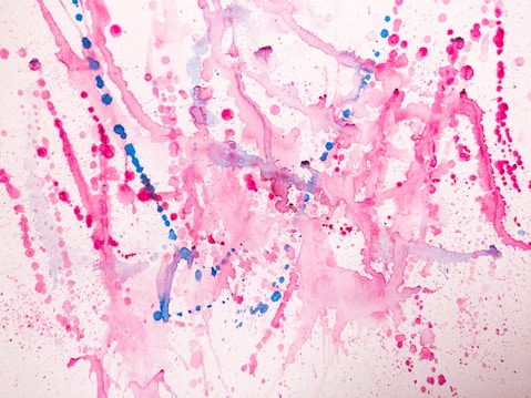Vibrant splashes of pink and blue paint create an abstract pattern on a white background. The composition consists of various dots, splatters, and fluid lines, with each color blending and overlapping in places. The overall effect is energetic and playful, with a sense of dynamic motion capture.