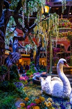 A vibrant indoor garden scene featuring a large artificial swan surrounded by colorful flowers and lush greenery. Towering trees with cascading foliage create a canopy, while a decorative fountain adds a touch of elegance to the setting. In the background, people can be seen near a well-lit commercial area with signage for stores or restaurants.