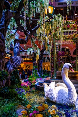 A vibrant indoor garden scene featuring a large artificial swan surrounded by colorful flowers and lush greenery. Towering trees with cascading foliage create a canopy, while a decorative fountain adds a touch of elegance to the setting. In the background, people can be seen near a well-lit commercial area with signage for stores or restaurants.