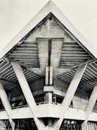 A striking geometric structure features large, angular beams supporting a concrete roof with an industrial design. The texture of the concrete is visible with strong lines and forms creating a sense of movement and architectural precision.