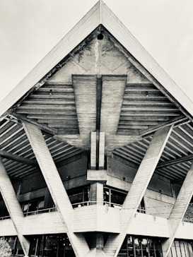 A striking geometric structure features large, angular beams supporting a concrete roof with an industrial design. The texture of the concrete is visible with strong lines and forms creating a sense of movement and architectural precision.