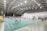 A spacious indoor badminton court with numerous people playing and observing games. The ceiling is high and covered with numerous lights, indicating a well-illuminated and possibly modern sports hall. Several courts are visible, separated by blue nets.