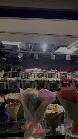 A flower shop display with various bouquets of flowers wrapped in pastel-colored paper. The background features a wall with numerous small bouquets arranged in rows, labeled with price tags. Overhead lighting illuminates the scene, casting a soft glow over the vibrant colors of the flowers.