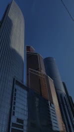 Skyscrapers with modern architectural designs rise against a clear blue sky. The buildings feature sleek glass facades with reflective surfaces that capture the surrounding light and create interesting patterns. Their heights and geometric shapes tower impressively.