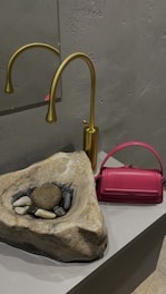 A modern bathroom setting features a stone sink with a natural, rugged look, containing several smooth stones and pebbles. A sleek, gold tap is mounted on the wall behind the sink. To the right, a vibrant pink handbag adds a pop of color against the neutral tones of the stone and metal.