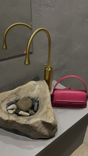 A modern bathroom setting features a stone sink with a natural, rugged look, containing several smooth stones and pebbles. A sleek, gold tap is mounted on the wall behind the sink. To the right, a vibrant pink handbag adds a pop of color against the neutral tones of the stone and metal.