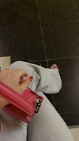 A person is holding a pink leather handbag while wearing light blue jeans. The person's nails are painted red, and they are wearing black sandals. The background consists of dark tiled flooring.