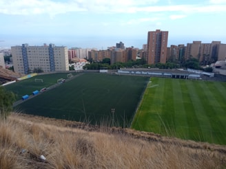 A large football field is situated amidst a cluster of tall apartment buildings. The field is divided into sections, with one area having artificial turf and another with natural grass. Goalposts are visible on both sides of the field. The surrounding buildings are multi-story and appear residential. Dry grass occupies the foreground, and the sea is faintly visible in the background.