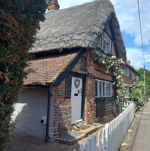 A charming, rustic cottage with a thatched roof, surrounded by climbing roses. The brick facade features diamond-patterned leaded windows. A small white picket fence lines the front of the property, and a pathway leads to a wooden door adorned with a heart-shaped ornament. The cottage is nestled in a tidy and picturesque village street.