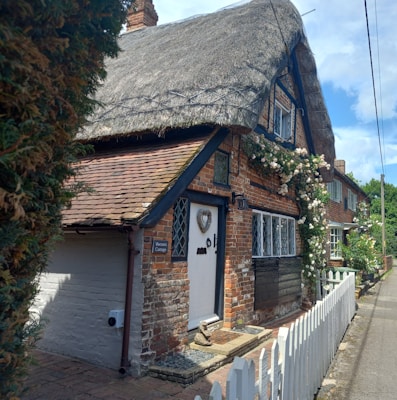 A charming, rustic cottage with a thatched roof, surrounded by climbing roses. The brick facade features diamond-patterned leaded windows. A small white picket fence lines the front of the property, and a pathway leads to a wooden door adorned with a heart-shaped ornament. The cottage is nestled in a tidy and picturesque village street.