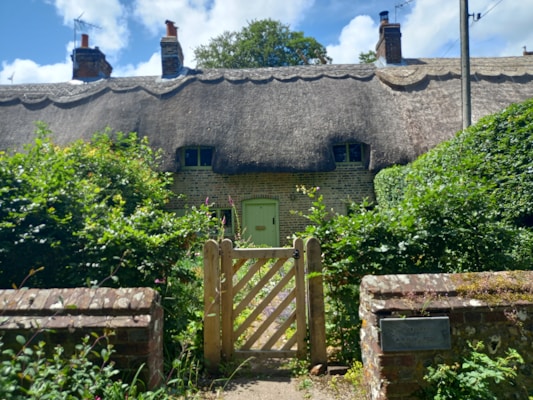 A charming cottage with a thatched roof is set amidst lush greenery and a blue sky. The brick structure is partially concealed by well-kept bushes and a wooden gate at the entrance. A brick pathway leads to the green front door, and chimneys can be seen rising from the roof, hinting at a cozy interior.