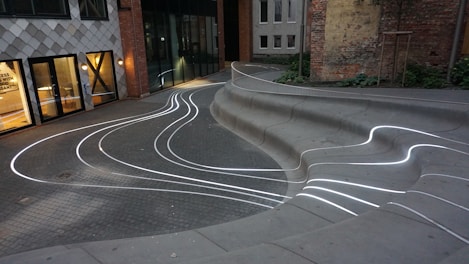 Curved concrete seating with embedded white LED lines creates a modern, artistic ambiance in an urban courtyard. The space is bordered by buildings with mixed architectural designs, including large windows and a checkerboard facade. Greenery and brick walls add contrast to the setting.